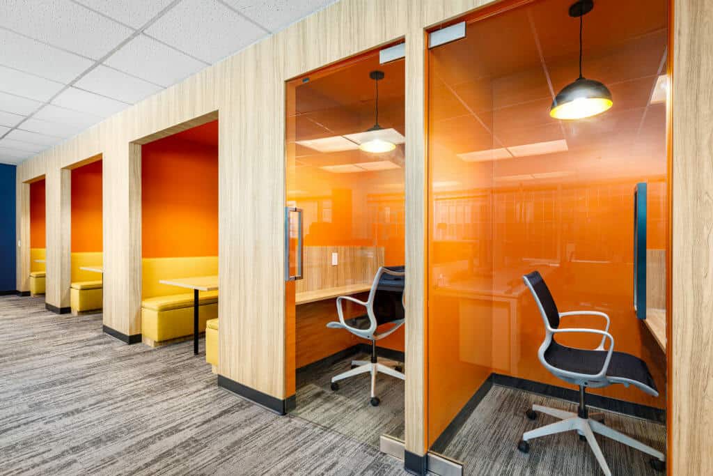 Une rang&eacute;e de cabines avec des portes vitr&eacute;es encastr&eacute;es dans un mur, une avec des bancs pour les r&eacute;unions et deux avec des chaises de travail et des bureaux