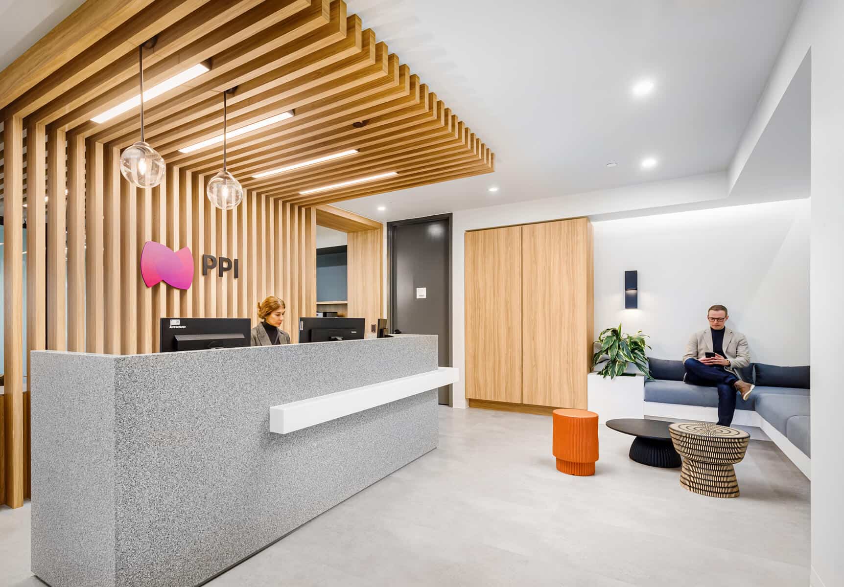 Espace de r&eacute;ception de bureau moderne avec un homme et une femme, des accents en bois &eacute;l&eacute;gants, un bureau d'entr&eacute;e, un coin salon et des suspensions suite &agrave; une r&eacute;novation de bureau.