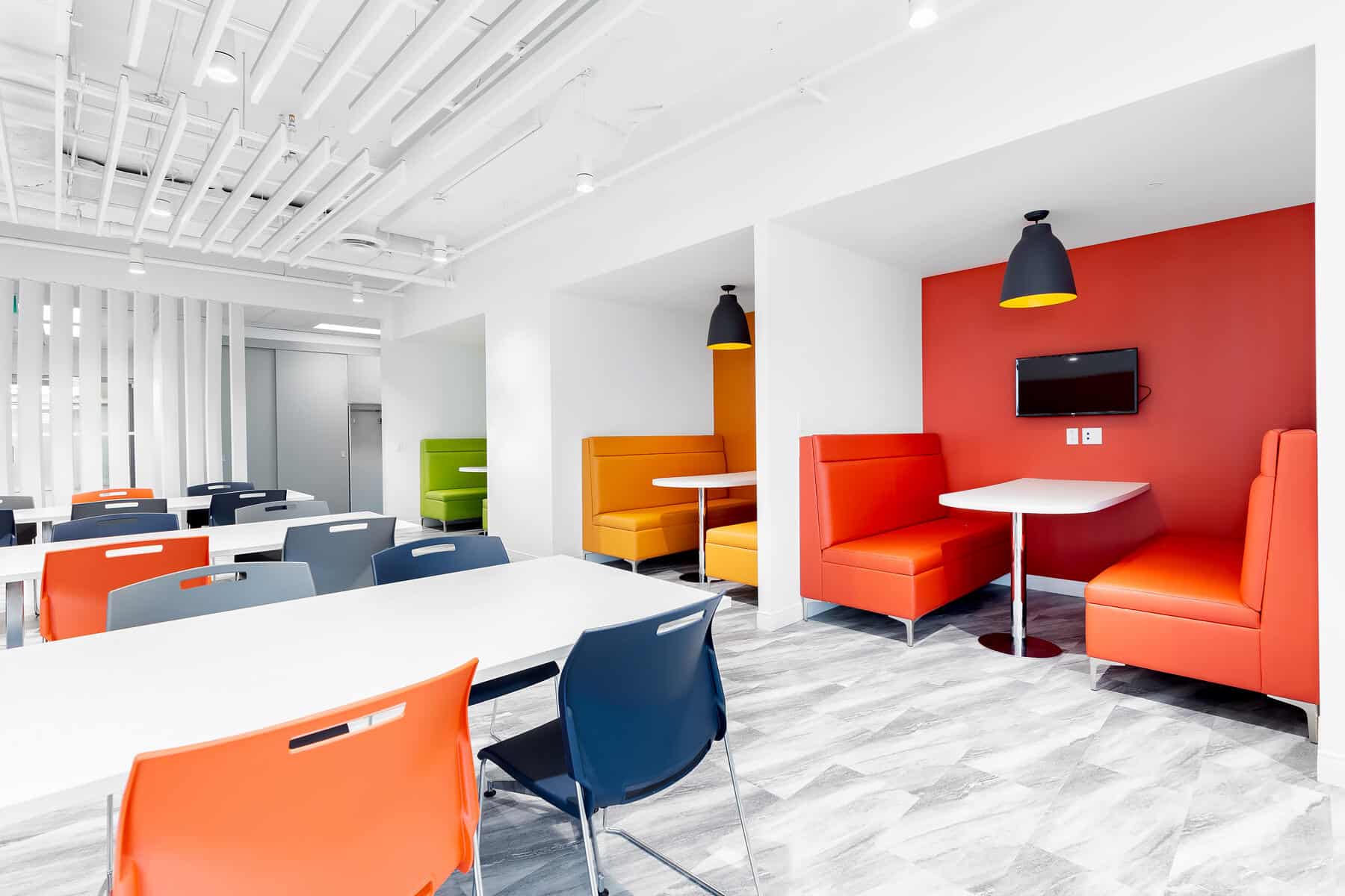 Salle de repos de bureau moderne de DUCA Financial avec des meubles color&eacute;s, notamment des cabines orange, des chaises vertes et orange et des tables blanches, sous un &eacute;clairage vif.